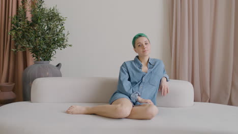 Young-Woman-With-Blue-Hair-And-Wearing-A-Denim-Shirt-Sitting-On-A-Big-Sofa-Looking-At-Side,-Then-Smiles-To-The-Camera