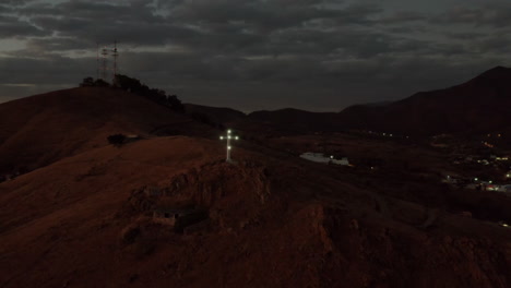 Solar-powered-Christian-cross-monument-on-hilltop-after-sunset,-aerial