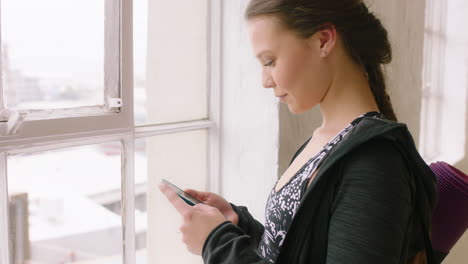 attractive yoga woman using smartphone texting sharing training practice on social media looking out window planning ahead in wellness studio