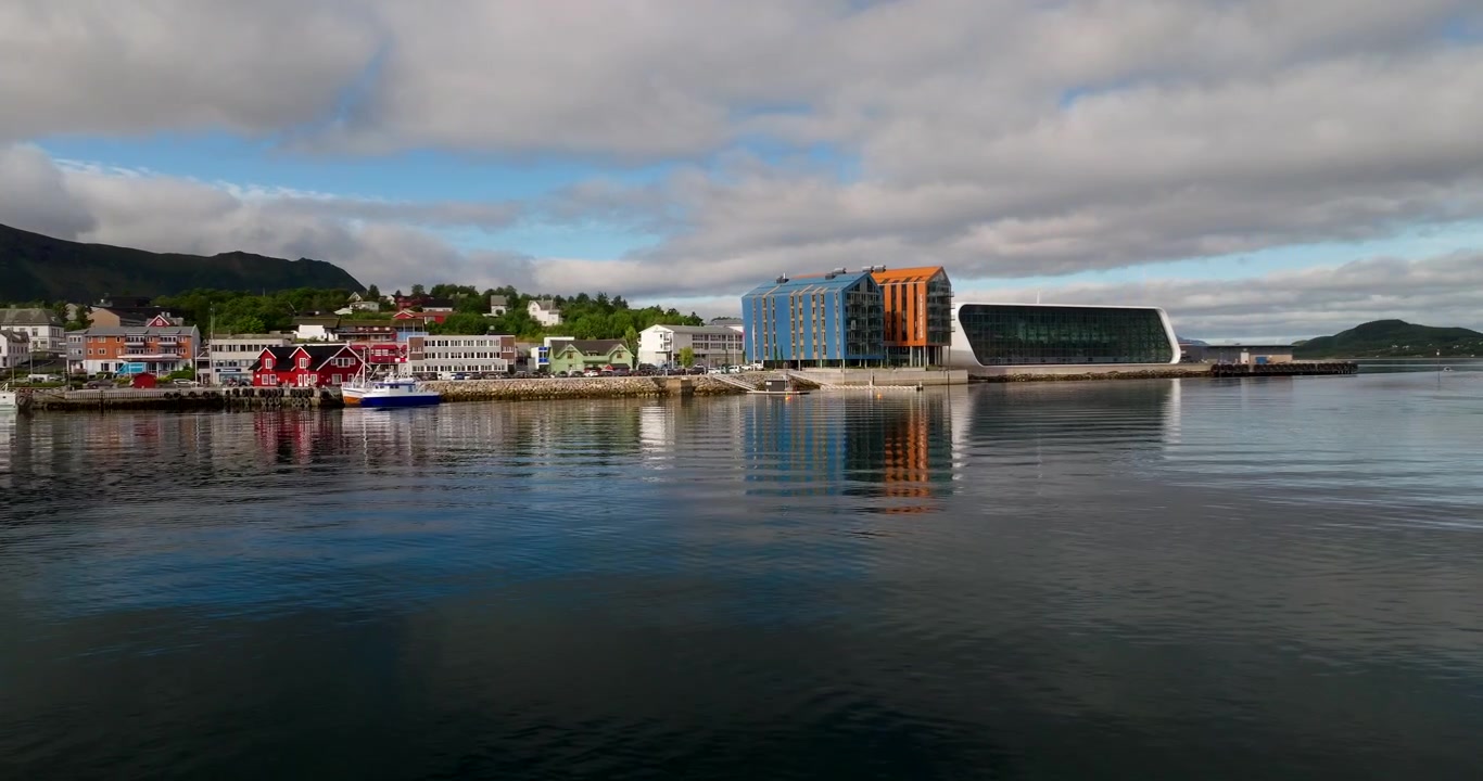 Etablerande drönarvy med dollyrörelse över Stokmarknes bro och hamn vid vattnet, speglade i lugnt vatten.