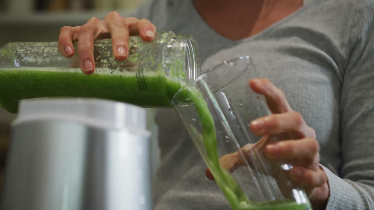 Běloška nalévá zelený zeleninový smoothie do skleněné nádoby v kuchyni.