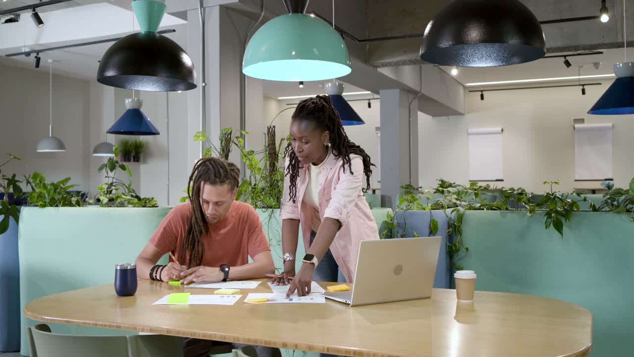 African American woman approaching and pointing, guiding seated man writing sticky notes by laptop