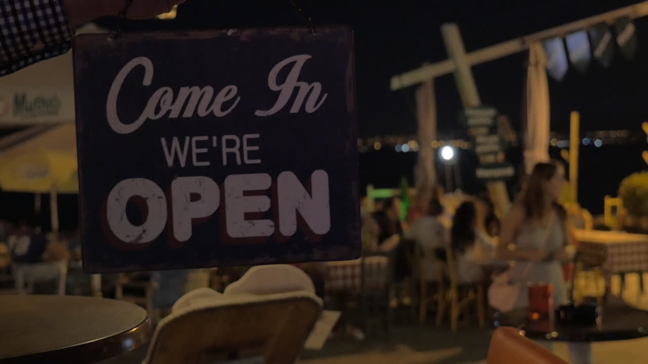 Insegna di un negozio che da 'chiuso' si gira su 'aperto' di notte, con lo sfondo di un caffè sulla spiaggia a Salonicco, Grecia