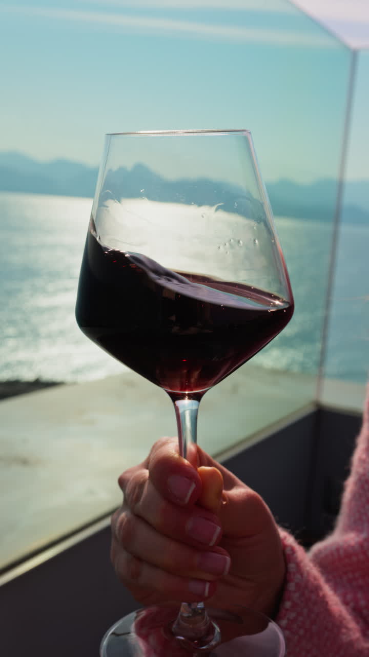 Close up of a woman swirling a glass of red wine on a table at a terrace with a sea view. Vertical