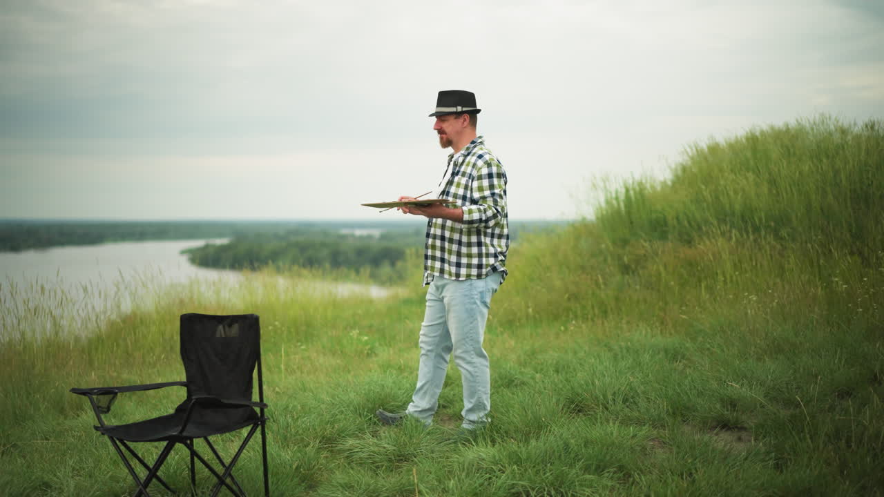 An artist in a checkered shirt, hat, and jeans holds a palette as he walks slowly in a lush grass field beside a serene lake. A black chair is visible in the background