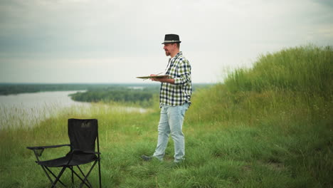 an artist in a checkered shirt, hat, and jeans holds a palette as he walks slowly in a lush grass field beside a serene lake. a black chair is visible in the background