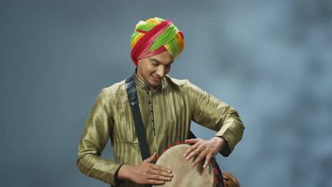 Portrait-of-young-cheerful-Indian-man-in-traditional-clothes-and-turban-playing-a-drummer-and-smiling-at-camera