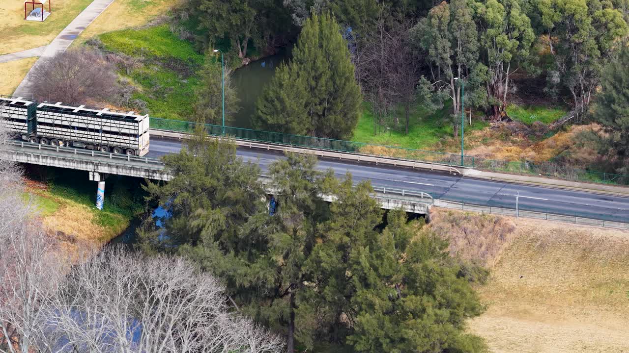 A livestock transport truck drives across a countryside bridge surrounded by trees and grassland, captured in daylight from an elevated aerial perspective