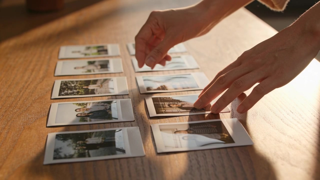 Hands in beige longsleeve lifting, steadying instant prints on wooden table aligning under light