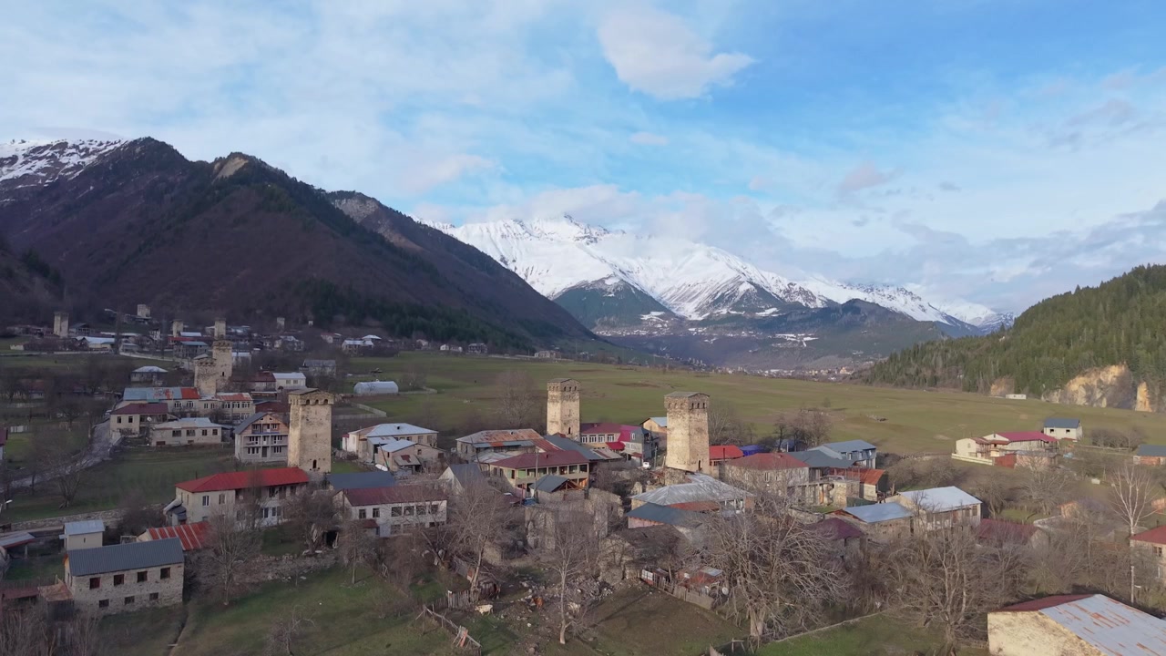 Drone subindo acima de Mestia, revelando casas tradicionais, icônicas torres Svan e as majestosas Montanhas do Cáucaso cobertas de neve sob um céu alpino claro