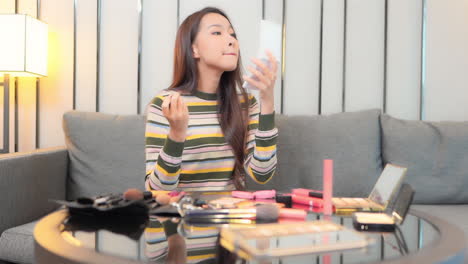 while sitting on a hotel suite couch, a pretty young woman looks in a hand mirror and applies lipstick