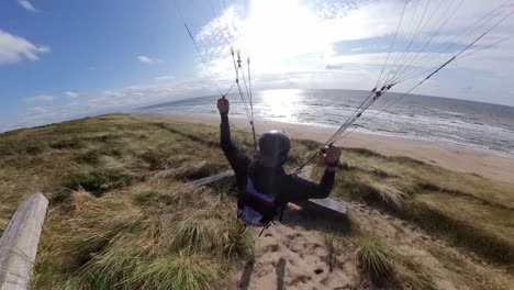 Close-up-following-shot-of-an-extreme-paraglider-skimming-over-the-dunes,-tapping-benches-and-zipping-over-the-grass-on-a-sunny-day