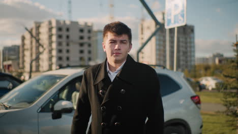 man in black coat exiting his car with a serious expression in vibrant urban area featuring parked cars, modern buildings, greenery, moving vehicles, and pedestrians