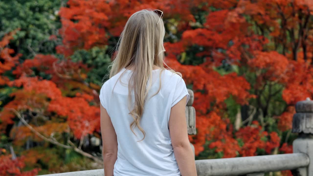A blonde girl strolls gracefully across a traditional bridge in Kyoto, surrounded by the breathtaking colors of autumn.