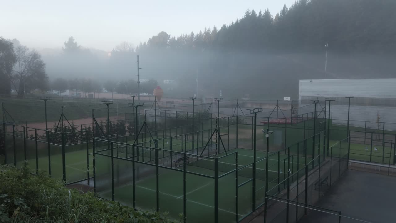 Dense morning fog gradually dissipating, revealing padel court nestled within verdant landscape of sant hilari sacalm, bathed in soft, emerging light of early autumn day
