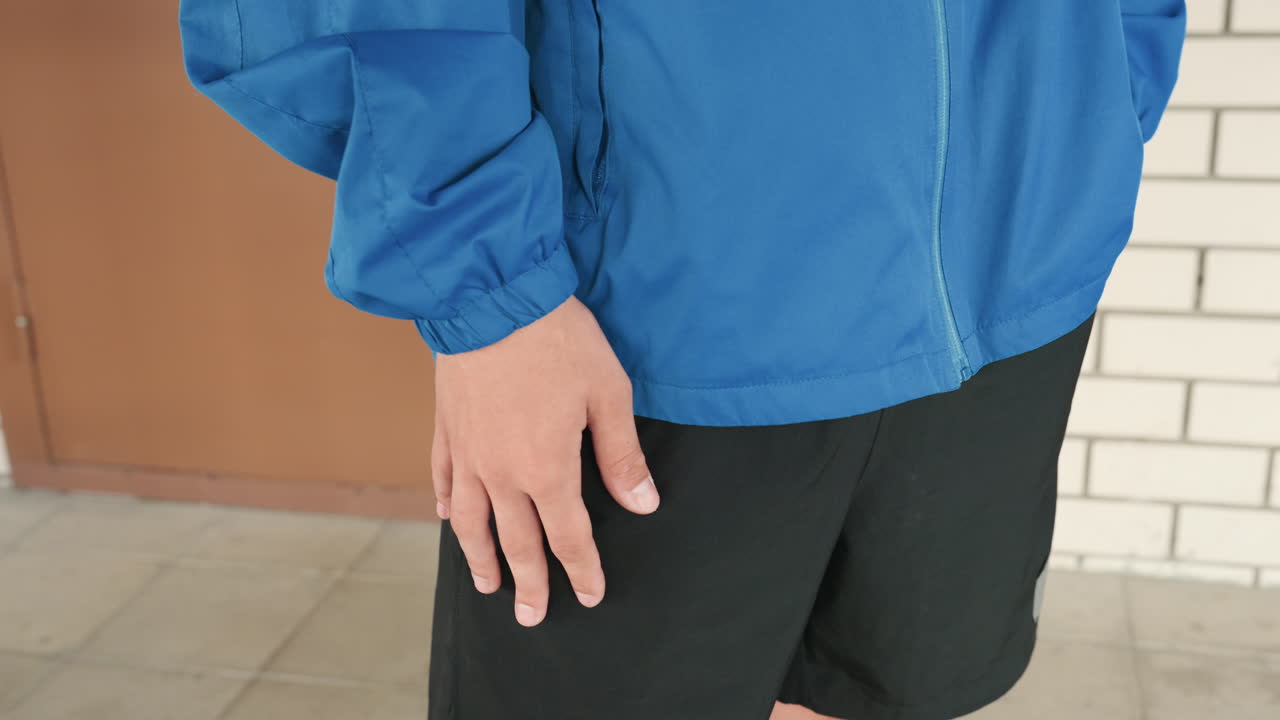นักกีฬาเยาวชนในเสื้อแจ็กเก็ตสีน้ำเงิน กำลังรออยู่ข้างทางเดินอิฐและม้านั่ง สวมกางเกงขาสั้นสีดำ มือปล่อยสบายๆ ข้างลำตัว โฟกัสและตั้งตารอก่อนเริ่มเกม รายละเอียดท่าทางและนิ้วที่เป็นธรรมชาติ ภายในอาคาร