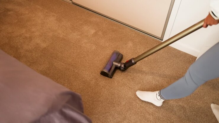 A woman uses a vacuum cleaner on a carpeted floor in a well-lit room, demonstrating cleaning action