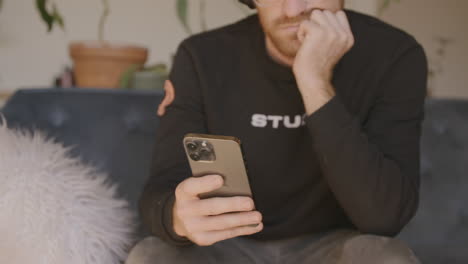 Medium-close-front-angle-of-a-man-sitting-on-a-couch-scrolling-and-swiping-and-typing-on-an-Iphone