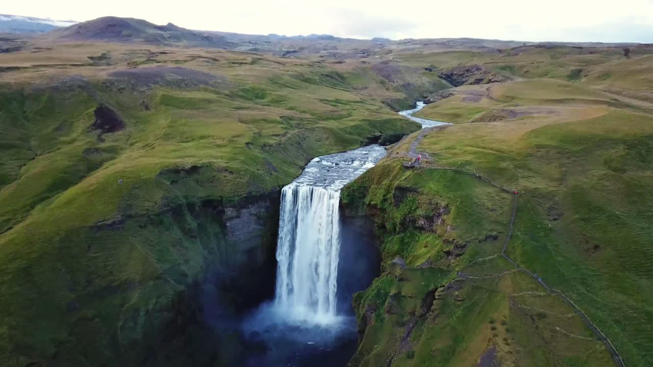 Breathtaking aerial view of Skógafoss, Iceland, with its majestic waterfall flowing over green cliffs into a misty pool. The serene landscape features lush meadows, rugged terrain, and a winding river