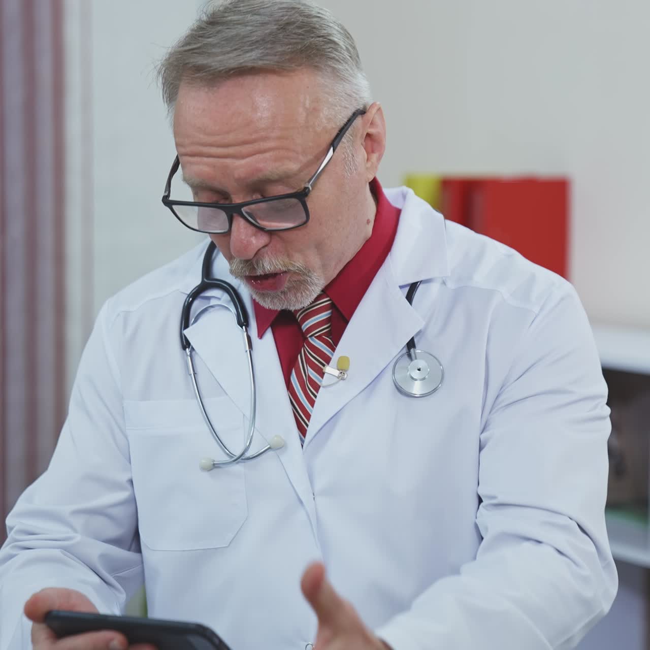 Medico maschio anziano seduto al tavolo del medico e che guarda il telefono. Medico esperto e pensieroso consulta il paziente tramite connessione video.