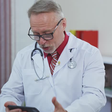 senior male doctor sitting at doctor's table and looking at phone. thoughtful experienced doctor consults patient via video connection