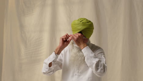 Studioaufnahme-Eines-älteren-Sikh-Mannes-Mit-Bart,-Der-Beim-Aufsetzen-Eines-Turbans-Vor-Einfachem-Hintergrund-Eine-Salai-Nadel-Verwendet-1