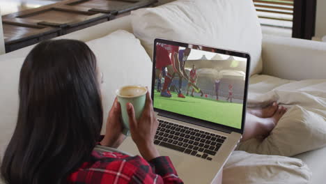 compuesto de una mujer sentada en casa sosteniendo un café viendo un partido de hockey en una computadora portátil