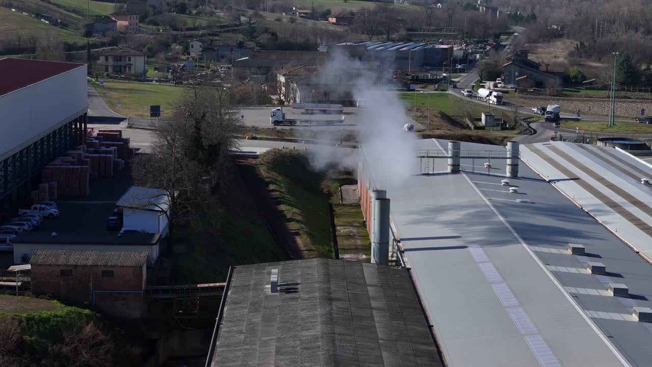 Veduta aerea di un impianto di produzione industriale che rilascia vapore bianco dalle ciminiere sul tetto, circondato da campagna rurale, terreni agricoli e piccoli edifici industriali in un paesaggio vallivo
