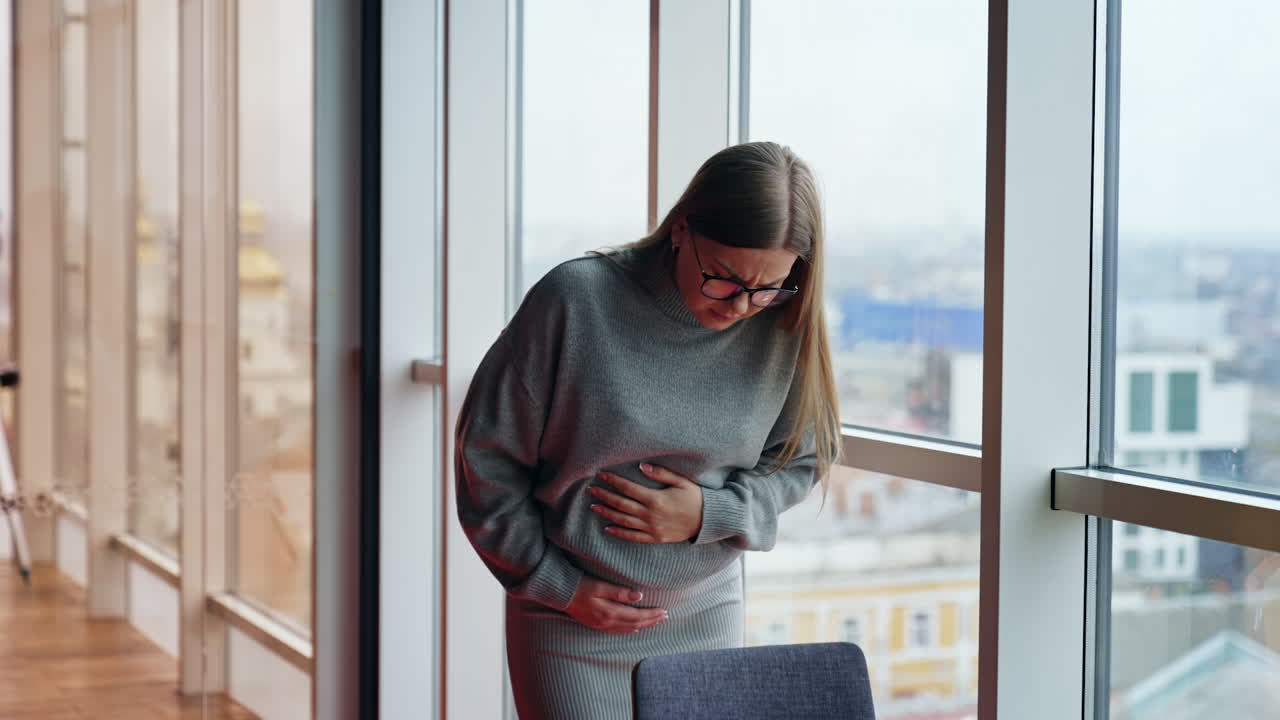 Pregnant woman suffering belly ache standing in the room with panoramic windows. Expecting mom feeling her baby kicking.