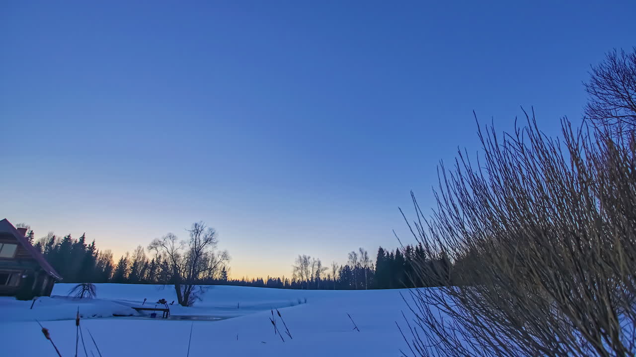 설원이 있는 추운 겨울날 푸른 하늘을 따라 나무 오두막 위로 아름다운 일출의 저속 촬영