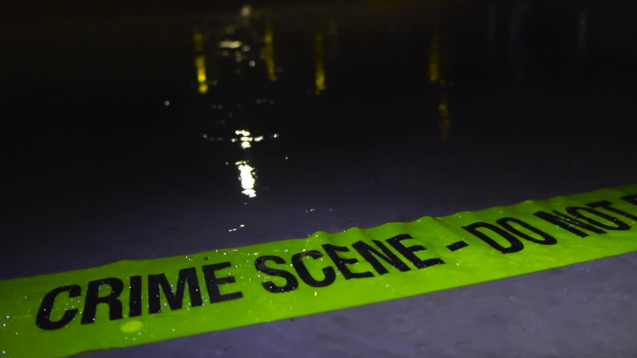 Close up or closeup footage of a yellow colored Crime Scene Do Not Enter police tape ribbon abandoned on a concrete water covered ground during a rainy night while raindrops fall in slow motion