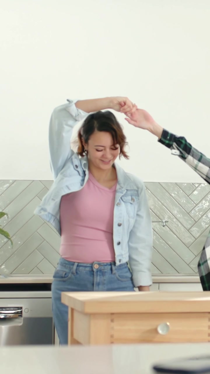 Vertical video: Smiling and holding hands two women twirling around island for fun in denim, plaid