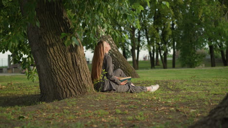 dama de ropa gris se sienta al aire libre bajo un árbol, leyendo un libro con la mano en la página, inclinando la cabeza y la pierna extendida, el fondo presenta vegetación y árboles