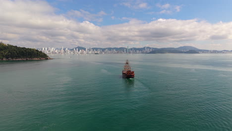Wide aerial shot features a pirate ship cruising across turquoise sea with the distant high-rise skyline and mountains of Balneário Camboriú, Brazil under partly cloudy skies