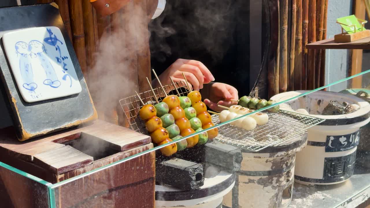 Primo piano di un venditore che griglia spiedini di mochi colorati su carboni ardenti con vapore che sale.