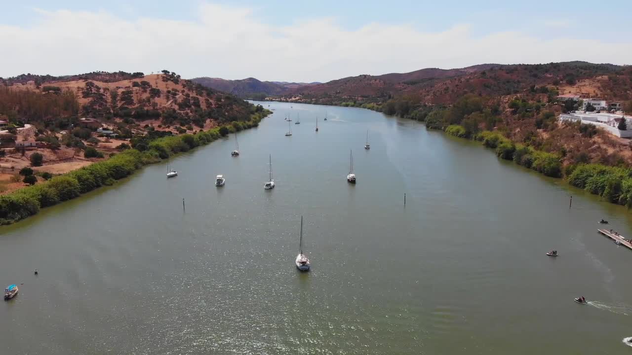 Aerial drone shot of riverside town in Alcoutim, Portugal, showing boats on water, white houses with red roofs, green hills, marina docks, peaceful atmosphere, and sunny weather