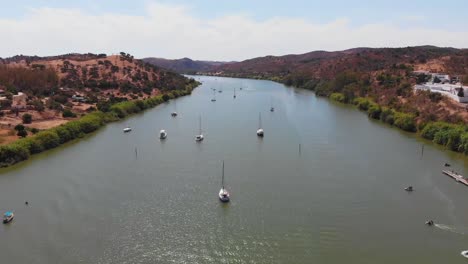 aerial drone shot of riverside town in alcoutim, portugal, showing boats on water, white houses with red roofs, green hills, marina docks, peaceful atmosphere, and sunny weather
