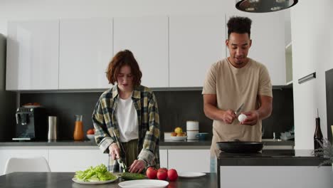 Happy-young-Black-brunette-man-cooking-scrambled-eggs-during-his-breakfast-preparation-along-with-his-mature-young-girlfriend-with-brown-hair-and-bob-hairstyle-in-the-kitchen-in-the-morning