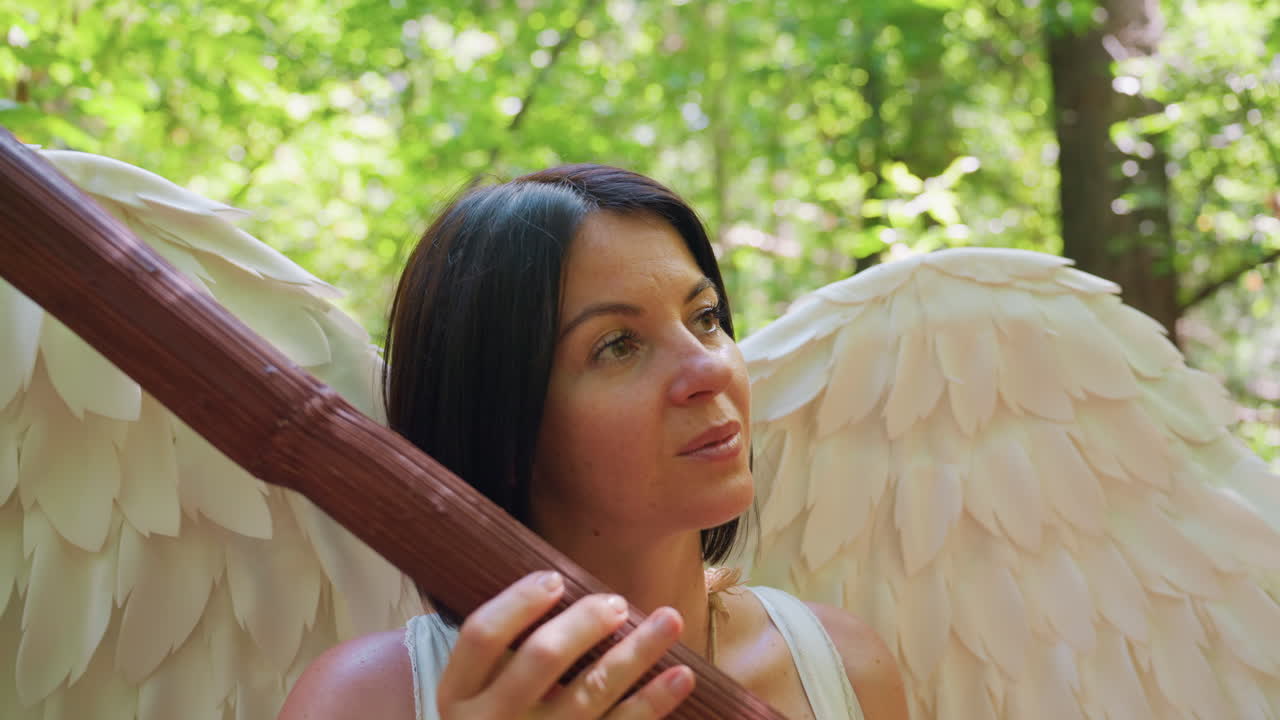 Dea fatata seduta nella foresta che tiene in mano un bastone di legno, picchiettandolo delicatamente come un'arpa mentre chiude gli occhi alla luce del sole, circondata da soffici foglie verdi e ali bianche, che esprime pace