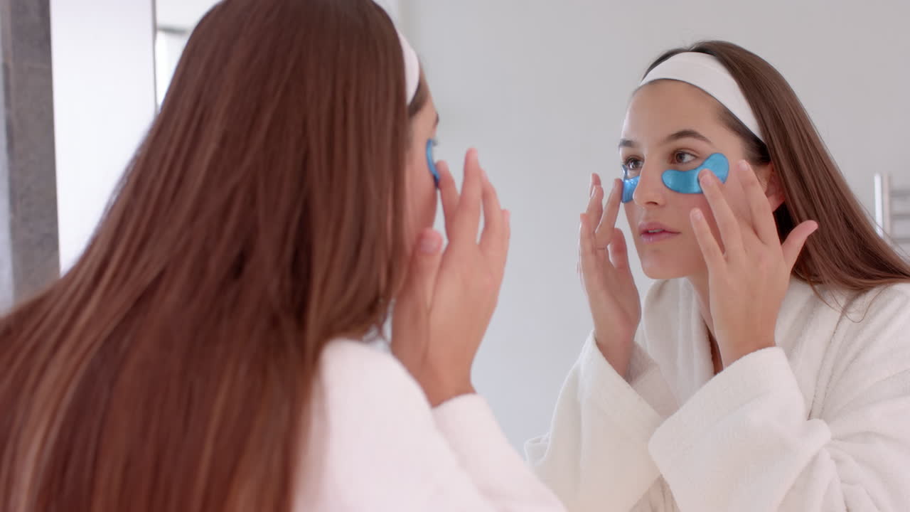 Applying blue eye patches, woman looking in mirror during home spa routine