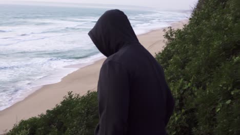 hooded person dressed in black going down a cliff, beach with waves in the background at sunset