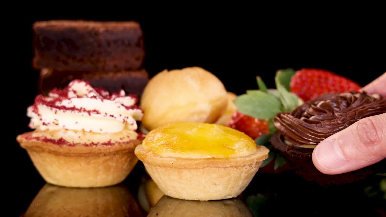 A hand picks up a chocolate brownie from a vibrant assortment of bakery desserts, including tarts, cream puffs, and strawberries, under bright studio lighting with a black background