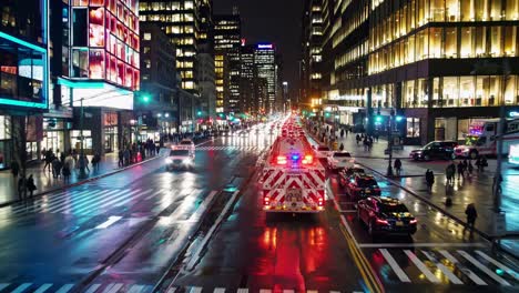 firetruck with flashing lights driving on a busy street in new york city at night, reflecting on wet asphalt with pedestrians walking on sidewalks and illuminated buildings in background