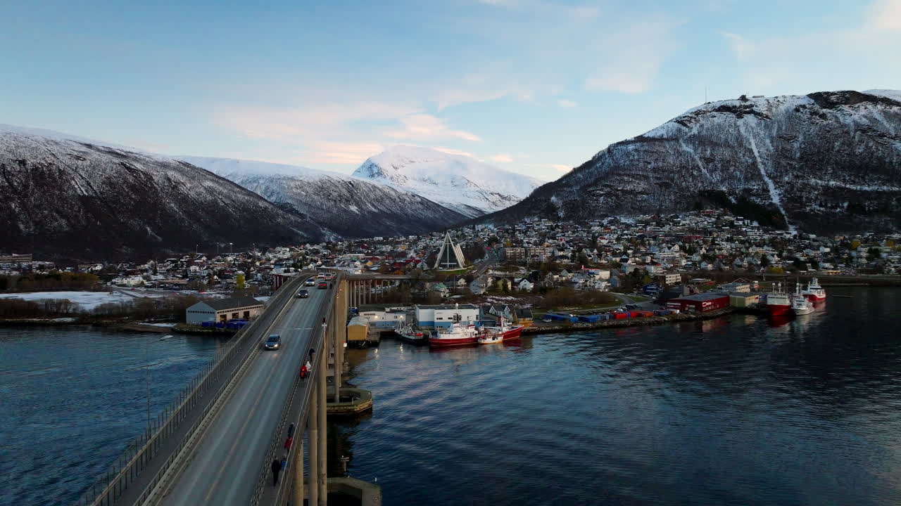 Kinoreife Drohnenansicht der Tromsø-Brücke und der Eismeerkathedrale in Norwegen im Winter. Luftaufnahme bei Dämmerung von Stadtverkehr und schneebedeckten Bergen.