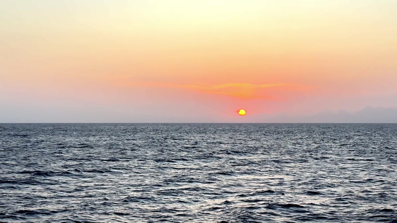 Vibrant crimson sun dips below the horizon of the vast open ocean during a serene summer evening captured from a low sea level perspective with soft hazy clouds and golden light across the water