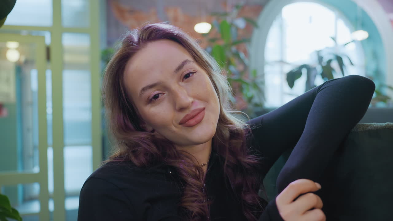 Relaxed woman with long wavy hair leans on cushion and smiles gently with closed eyes in softly lit interior surrounded by green plants and decorative elements in blurred background