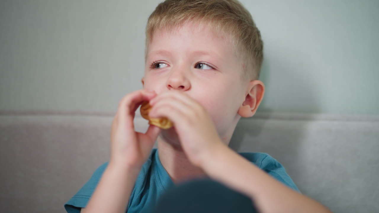 Portrait view of young boy wearing blue shirt smiling softly while eating sausage roll and looking at someone indoors capturing joyful casual moment of childhood snack time