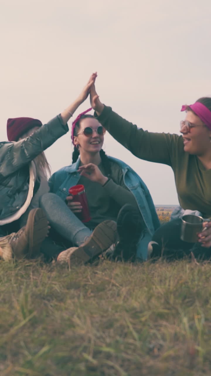 cheerful girl friend tourists with cups rest talking on green grass by tent against clear evening sky low angle shot
