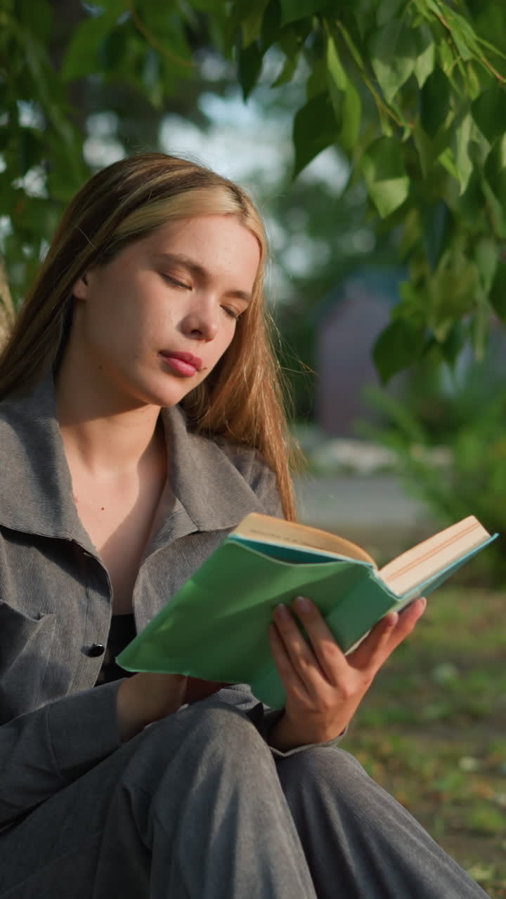 dama con ropa gris sentada al aire libre hojeando un libro con la cabeza inclinada ligeramente a la derecha, mirando pensativa, el fondo presenta vegetación y edificio borroso