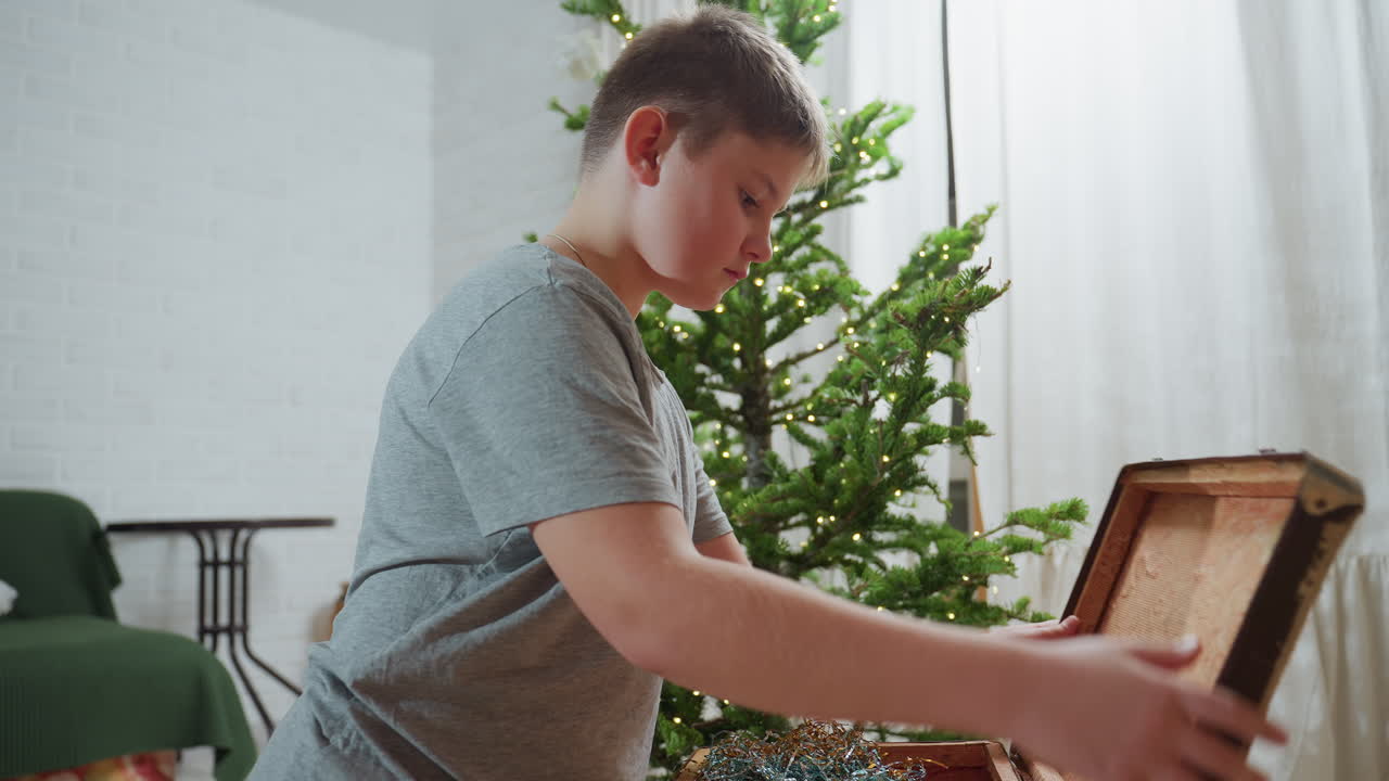 Boy in grey top calmly opening vintage box filled with colorful ornaments in bright cozy room decorated with Christmas tree and twinkling fairy lights, focused on finding beautiful holiday decoration